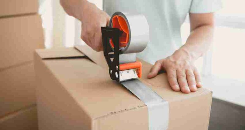 A person seals a cardboard box with gray packing tape using a tape dispenser. Other boxes are visible in the background, suggesting a moving or packing process.