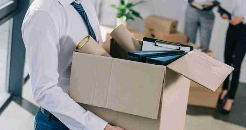 A person in business attire holds a cardboard box filled with office items, suggesting they are packing or moving. Another person is seen blurred in the background, also holding folders.
