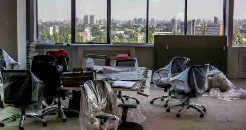 An office with swivel chairs covered in plastic, a table with items on it, and large windows showing a cityscape in the background. The office appears empty and unfinished.