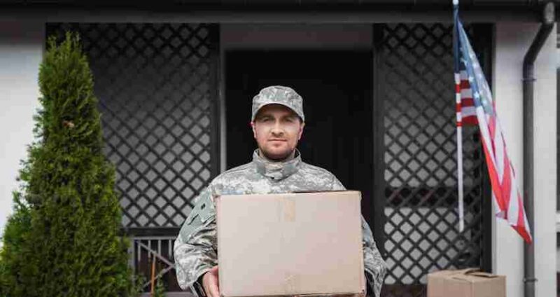 A person in military uniform holds a cardboard box while standing in front of a house with an American flag and a small green bush visible.