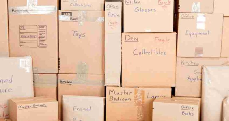 Stacked cardboard moving boxes labeled with contents like Toys, Glasses, Collectibles, and Kitchen, showing an organized moving or storage process.