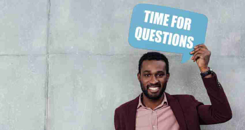 A smiling man in a suit holds up a blue sign shaped like a speech bubble that says, TIME FOR QUESTIONS, while standing against a gray wall.