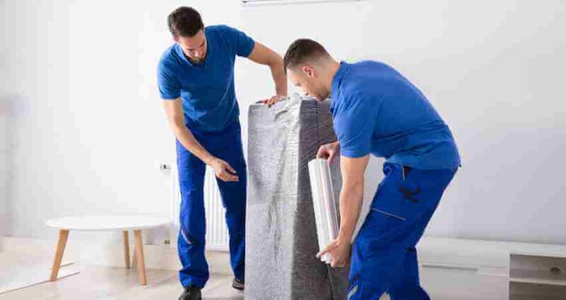 Two men in blue uniforms wrap a piece of furniture with protective padding in a bright, minimalistic room with white walls and light wood flooring.