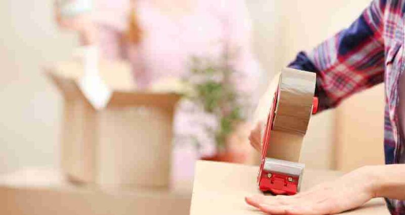 Close-up of a person sealing a cardboard box with packing tape. In the background, another person is unpacking a box with a small potted plant. Both are indoors, preparing for a move.