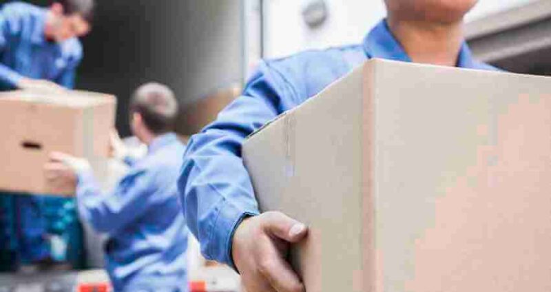 Two people in blue uniforms load cardboard boxes into the back of a truck. The focus is on one person carrying a box in the foreground, while another person is seen in the background with a box.