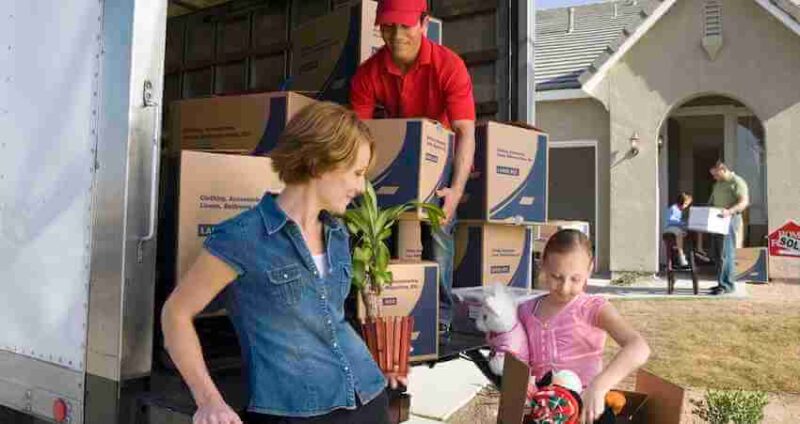 A woman and girl unload items from a moving truck, while a man in a red shirt carries a box. Cardboard boxes are stacked in the truck. A house with a sold sign is in the background.