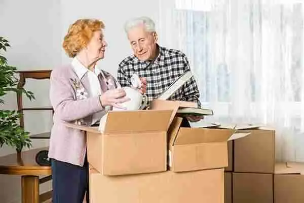An elderly couple stands among moving boxes, smiling as they unpack household items, including a vase and books, in a bright room with white curtains.