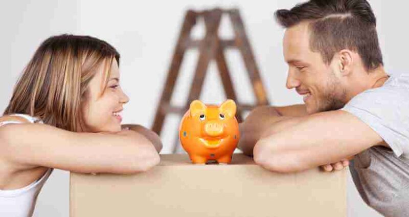 A woman and a man smile at each other while leaning on a box with an orange piggy bank between them. A ladder is visible in the background.