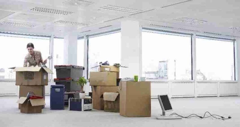 A person unpacks boxes in a large, empty office with big windows. Various boxes, a plant, and office equipment are scattered on the floor.