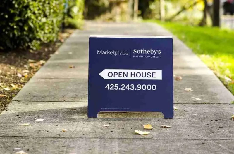 A blue sandwich-board sign on a sidewalk reads, Marketplace Sothebys International Realty, Open House, 425.243.9000. Trees and greenery are visible in the background.