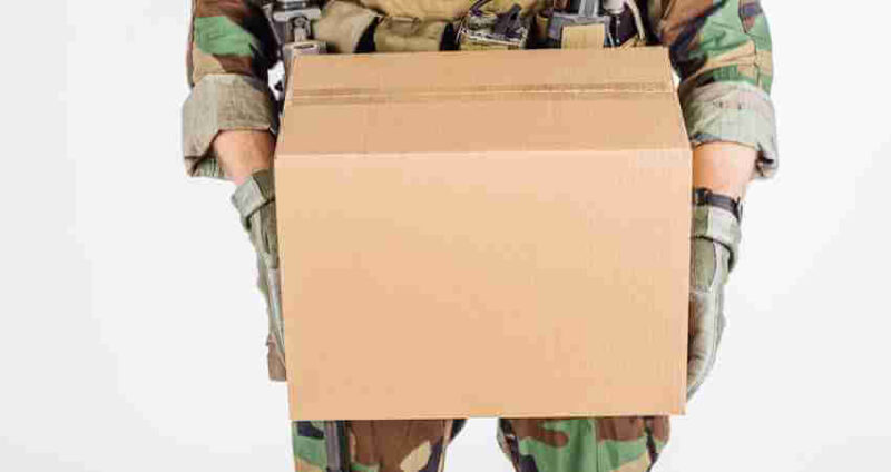 A person in camouflage military uniform holds a plain cardboard box against a white background.