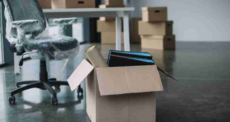 An office with a wrapped chair, a desk, and several cardboard boxes. One open box in the foreground contains rolled paper and binders, suggesting an office move or relocation.