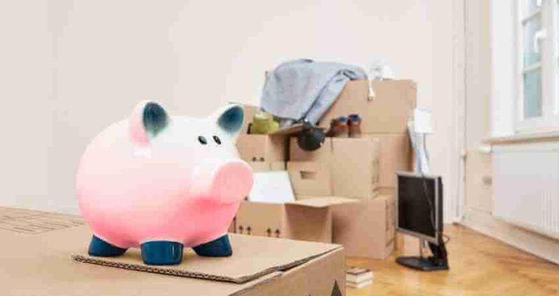 A pink piggy bank sits on a cardboard box in the foreground, with several packed moving boxes and household items in a bright room in the background.