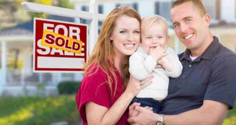 Smiling couple holding a young child stands in front of a house with a For Sale sign labeled Sold..
