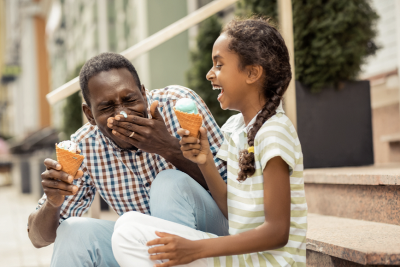A man and a girl sit on outdoor steps, laughing together while holding ice cream cones. The man covers his mouth, and the girl looks at him, smiling brightly. They appear to be enjoying a fun moment.