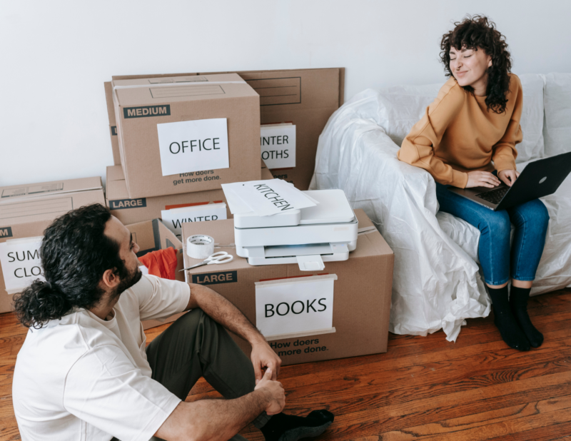 Two people talk in a living room with labeled moving boxes, a printer, and a laptop. The woman sits on a covered couch; the man sits on the floor. The floor is wooden, and the boxes are labeled by room and item.