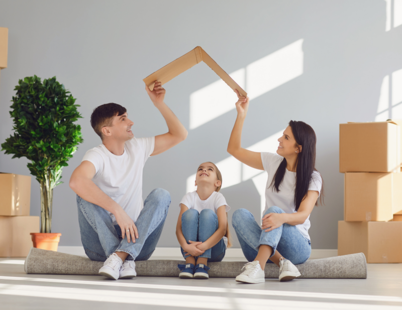 A family of three sits on the floor with cardboard boxes around them. The parents hold a cardboard roof shape over their daughters head, symbolizing a new home. All are smiling and wearing white shirts and jeans.