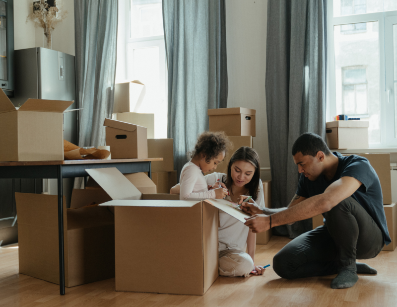 A family of three sits on the floor surrounded by moving boxes. The child sits inside a box, while the adults help her draw on it. Light streams in through large windows behind them.