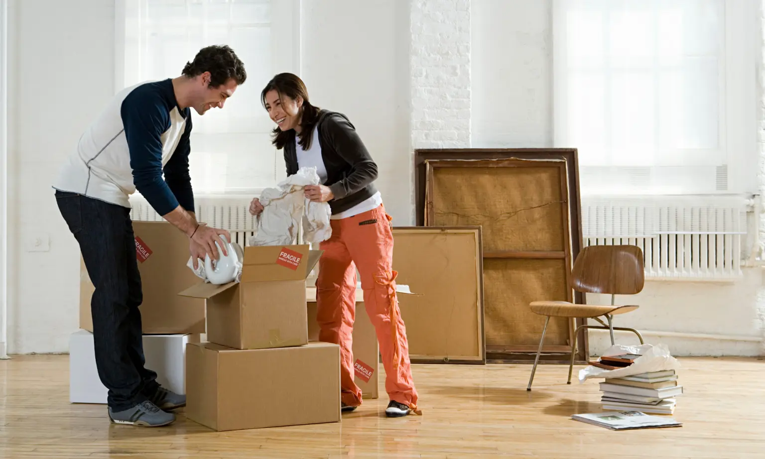 A man and woman unpack boxes in a bright room with large windows, art frames, and scattered furniture. They smile while handling dishes, suggesting they are moving in or settling into a new home.
