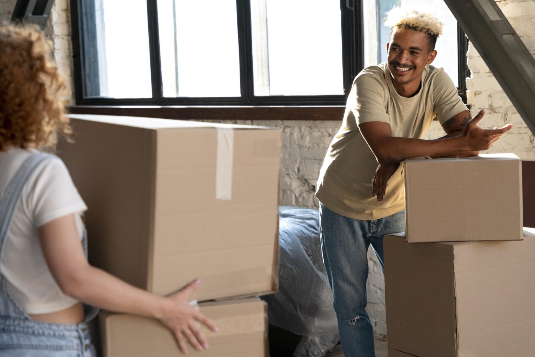 Two people smile and talk while unpacking cardboard boxes in a sunlit room with a large window, suggesting they are moving in or organizing a new home.