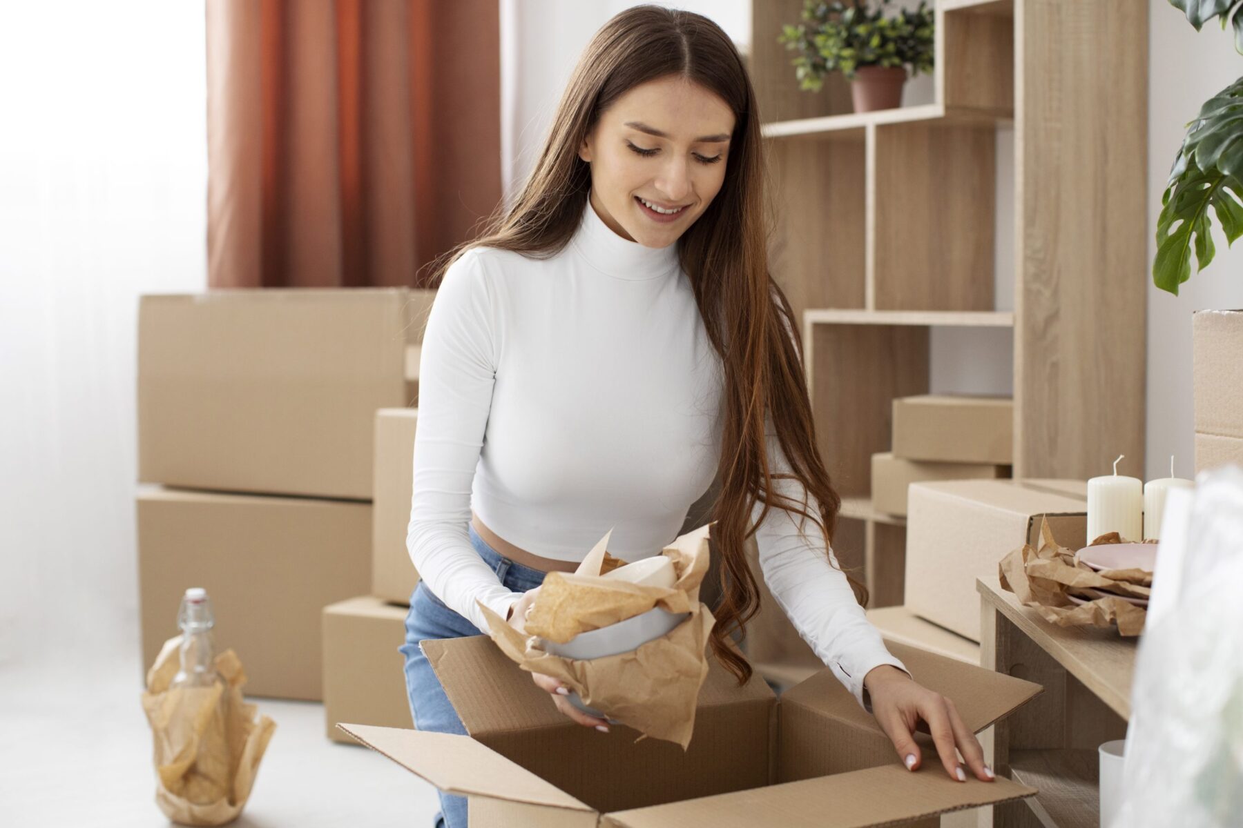 A woman with long hair packs dishes into a cardboard box, surrounded by moving boxes and household items, in a bright room with shelves and a plant.