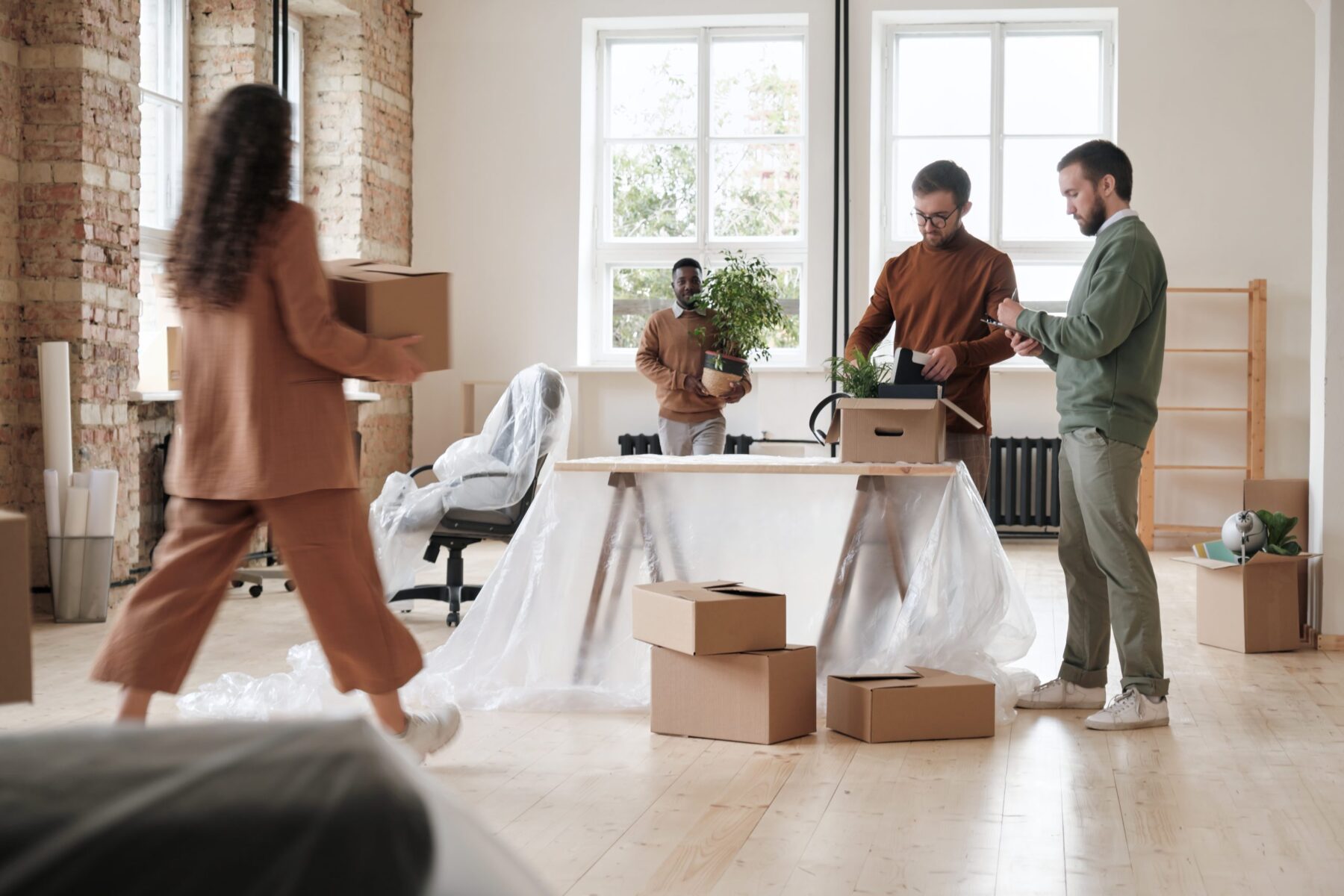 Four people are packing boxes in a bright room with large windows and brick walls. Some furniture is covered in plastic, and several cardboard boxes are scattered on the floor.
