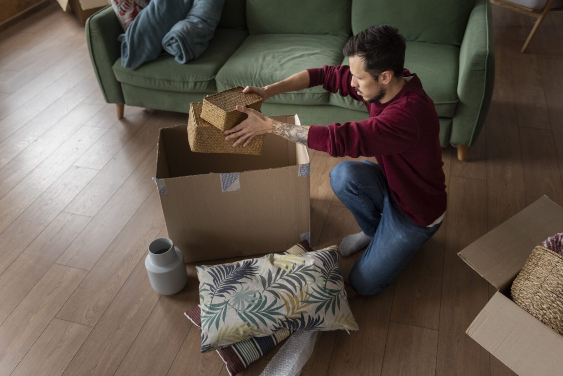 A person kneels on a wooden floor, packing a wicker box into a large cardboard box. Cushions, a vase, and another box are nearby. A green sofa is in the background.