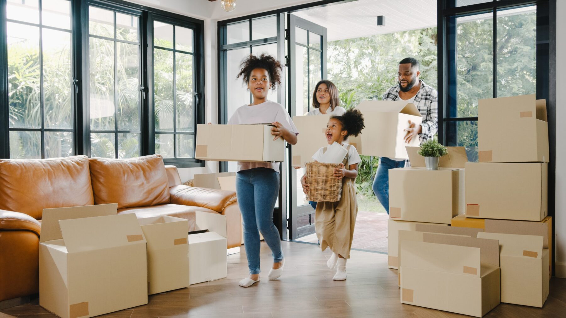 A smiling family of four carries moving boxes into a bright living room filled with packed cardboard boxes and a brown leather sofa.