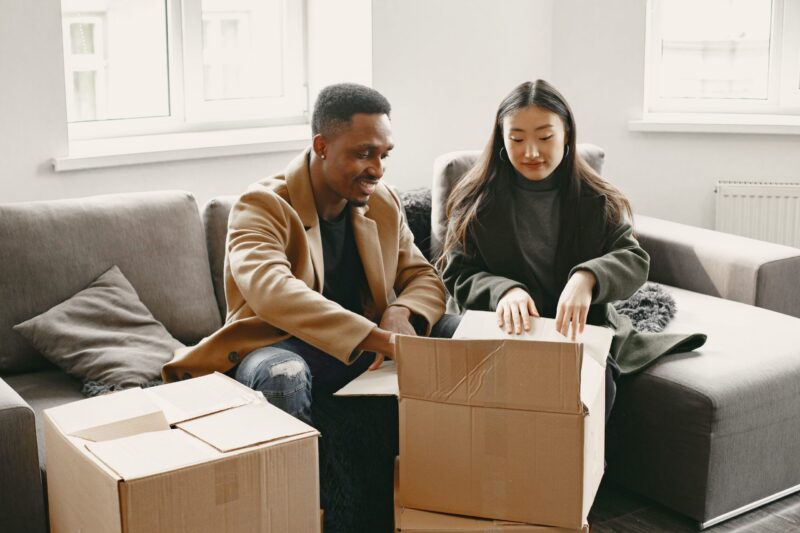 Two people sit on a gray couch in a bright living room, unpacking cardboard boxes together. They appear focused and content, possibly moving into a new home.