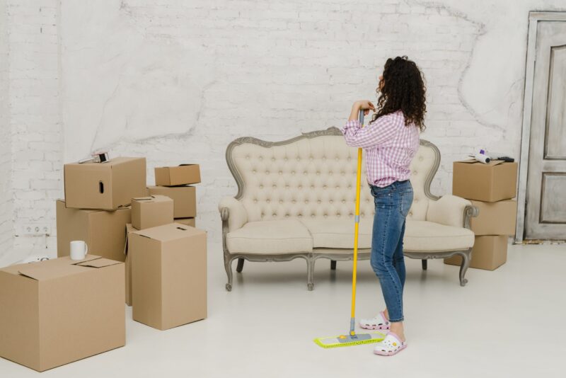 A woman stands with a mop in a room filled with cardboard boxes, in front of a vintage sofa and a white brick wall, suggesting she is cleaning before or after moving.