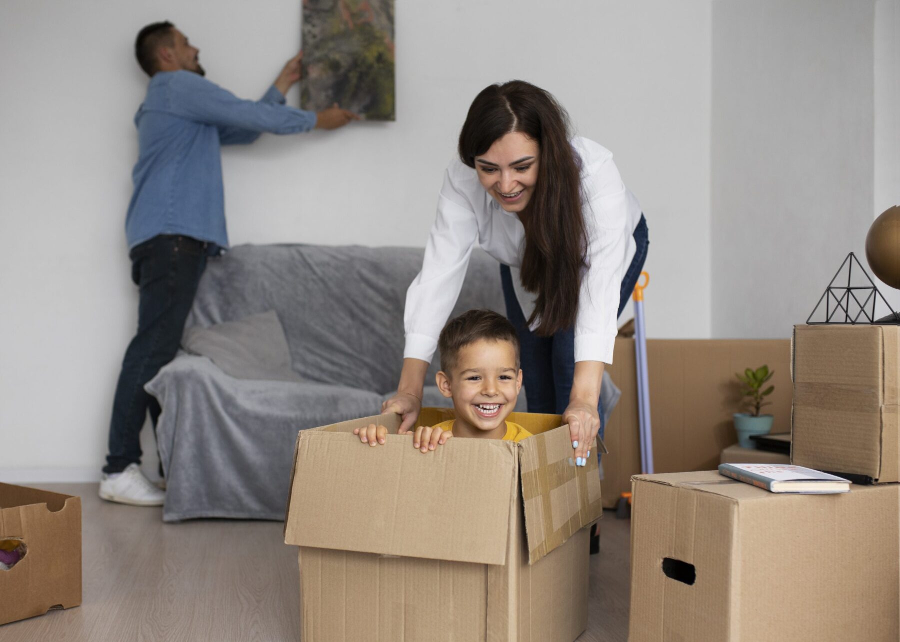 A woman pushes a smiling boy inside a cardboard box while a man hangs a picture on the wall in a living room filled with moving boxes.