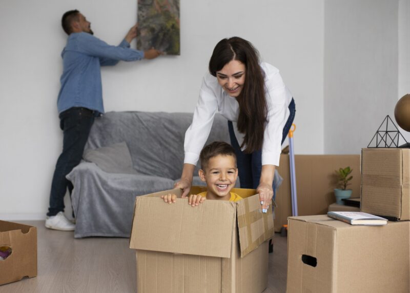 A woman pushes a smiling boy inside a cardboard box while a man hangs a picture on the wall in a living room filled with moving boxes.