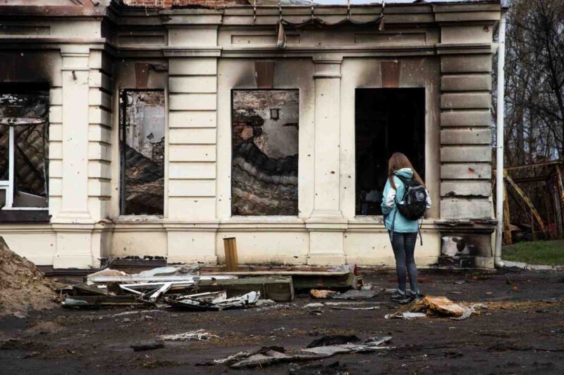 A person with a backpack stands in front of a damaged, abandoned building with broken windows, scattered debris, and signs of destruction.