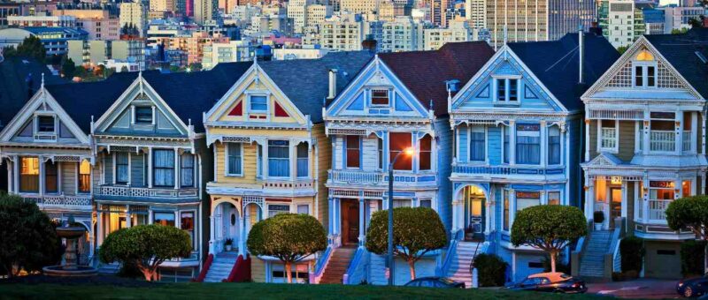Colorful Victorian-style houses in a row, known as the Painted Ladies, stand in front of a cityscape with tall buildings. Trees and parkland are in the foreground. The photo is taken at dusk with soft lighting.