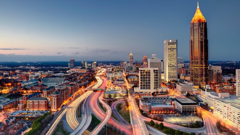 A cityscape at dusk shows illuminated skyscrapers and light trails from cars on highways winding through the urban area under a clear evening sky.