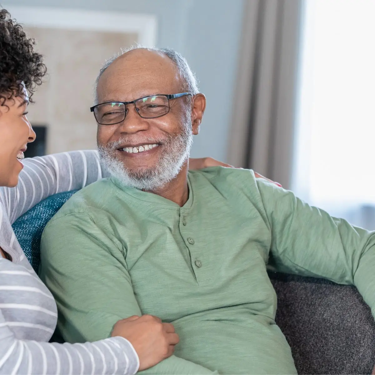 An older man with glasses and a gray beard smiles while sitting on a couch, looking at a woman next to him who is also smiling. They appear to be enjoying a pleasant conversation together.