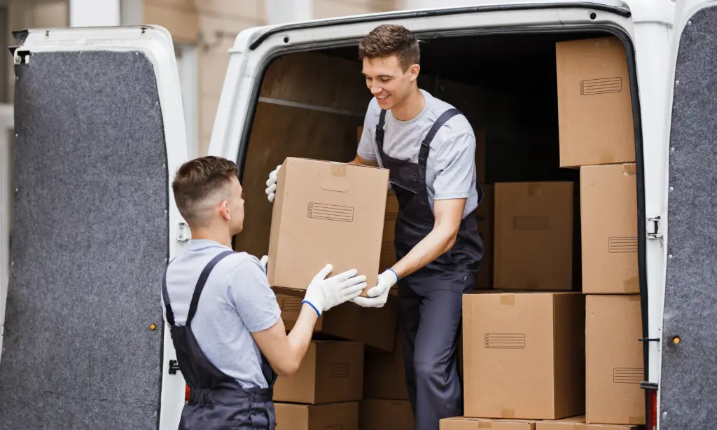 Two workers in uniforms unload cardboard boxes from a van. One stands inside handing a box to the other, who stands outside. The van is filled with many packed boxes.