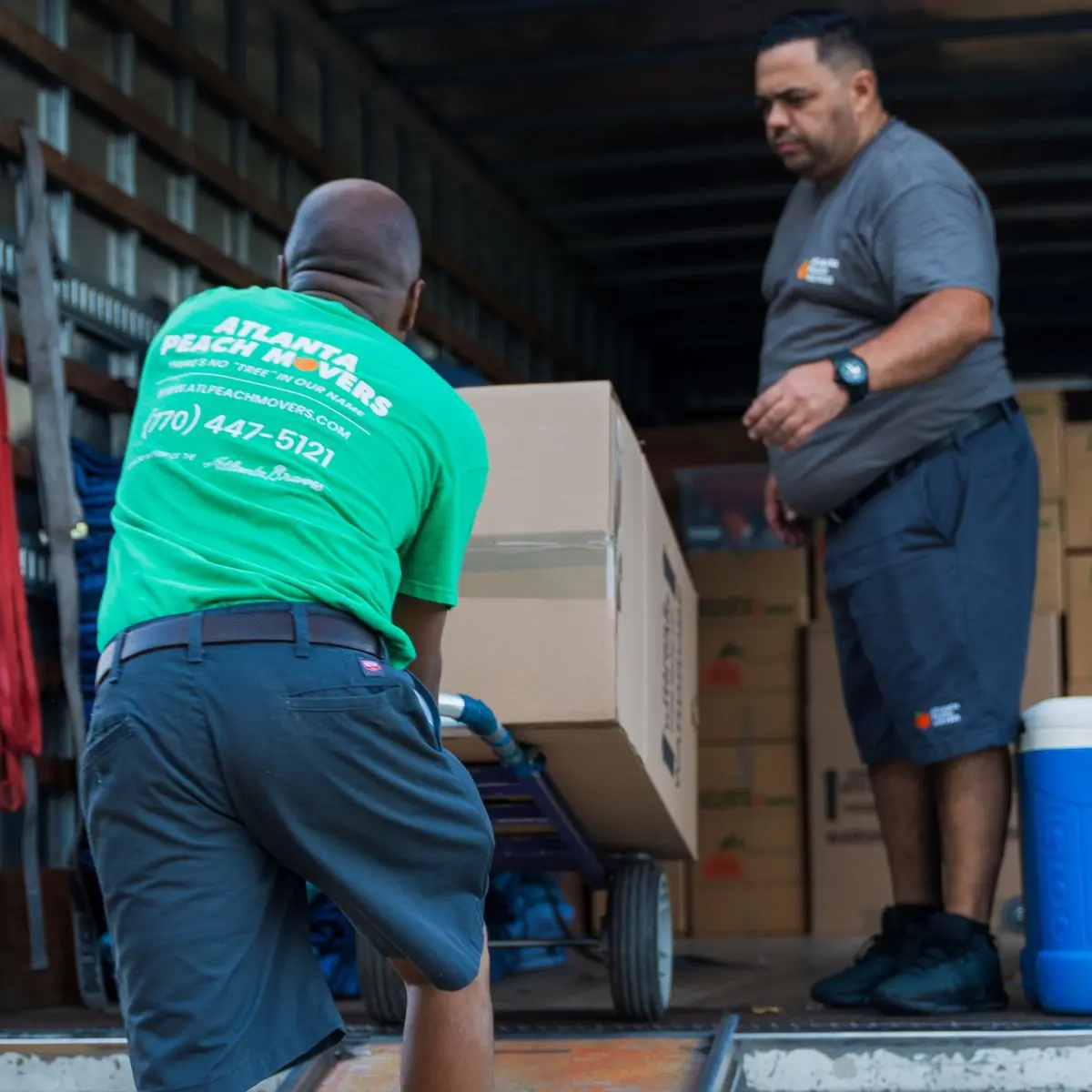 Two movers load boxes onto a dolly inside a moving truck. One wears a green shirt with business info, and the other wears a gray shirt. Stacked boxes and a blue cooler are visible in the background.