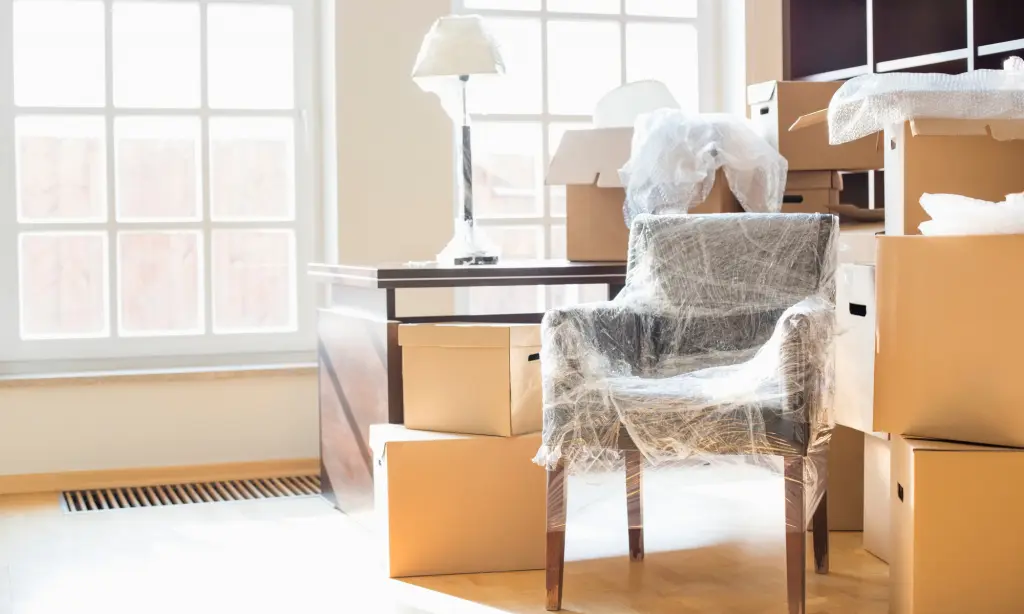 A room filled with moving boxes, a chair wrapped in plastic, a lamp, and a desk, with sunlight streaming through large windows in the background.