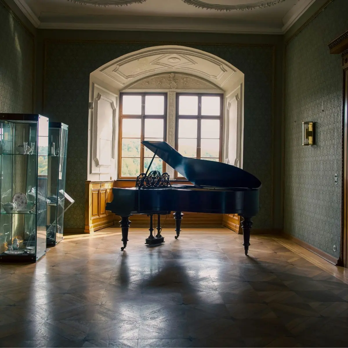 A grand piano sits in an elegant room with wooden floors, a large window letting in natural light, and a glass display cabinet filled with decorative items.