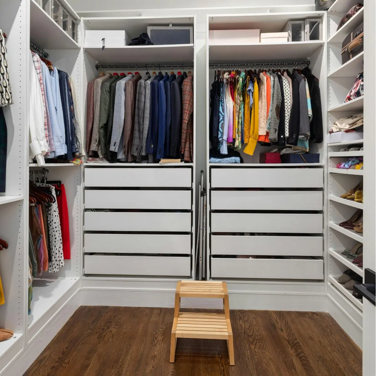 A neatly organized walk-in closet with clothes hanging, folded items, shelves with shoes, six drawers, and a wooden step stool on a wooden floor.