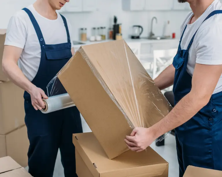 Two workers in overalls wrap a large cardboard box with plastic film in a bright, modern kitchen. Other cardboard boxes are stacked nearby, suggesting moving or packing activity.