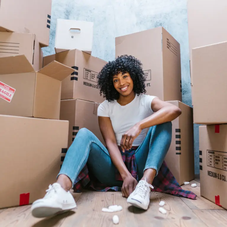 A woman in a white shirt and jeans sits smiling on a wooden floor, surrounded by stacked cardboard moving boxes and packing peanuts.