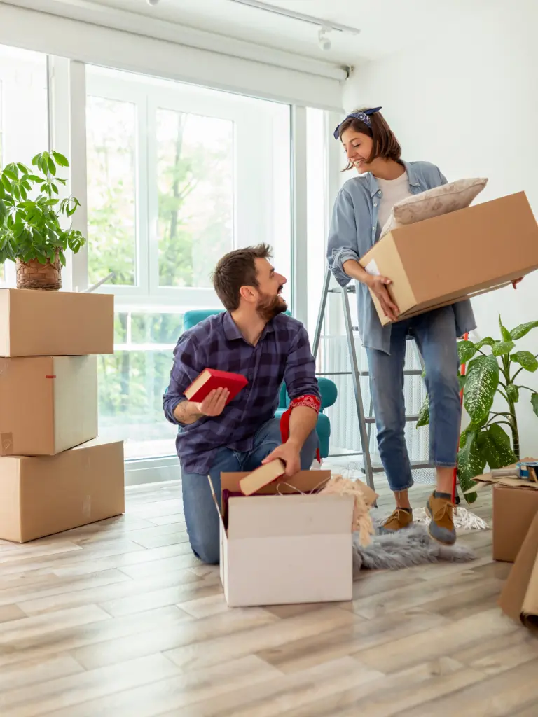 A man kneels on the floor holding a book, while a woman stands holding a box. They are surrounded by moving boxes and plants in a bright room with large windows.
