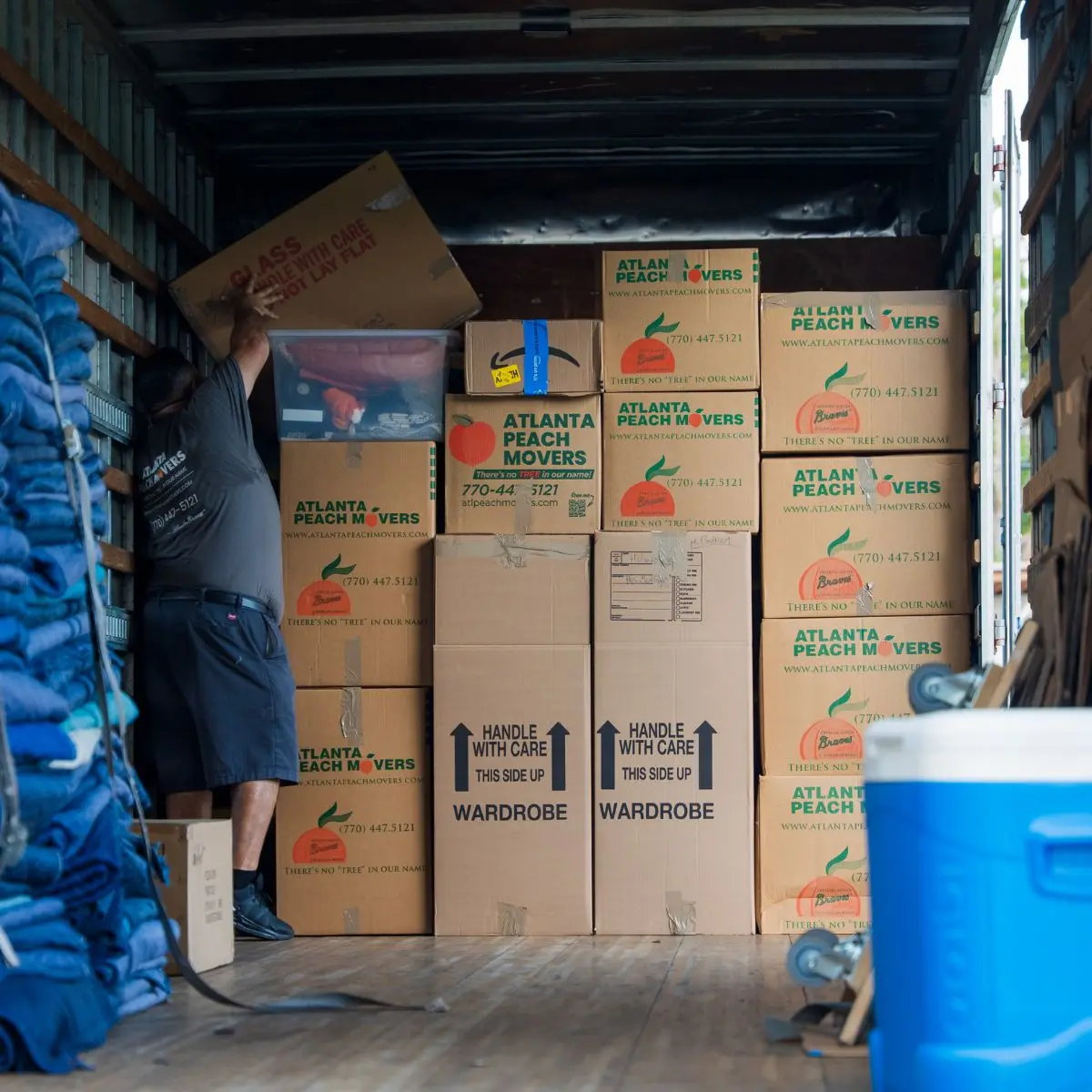 A person stacks a box inside a moving truck filled with labeled Atlanta Peach Movers boxes. A blue cooler and moving blankets are also visible in the foreground.