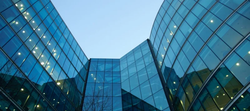 Modern glass office buildings with reflective windows curve toward each other, creating a geometric pattern against a clear blue sky. Lights inside the buildings are visible, adding a warm glow to the scene.