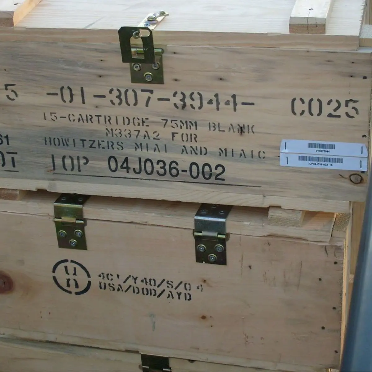 Wooden crate with black stenciled text indicating it contains 75mm blank cartridges for howitzers. The crate has metal clasps, latches, and a barcode label on the right side. Another crate is partially visible below.