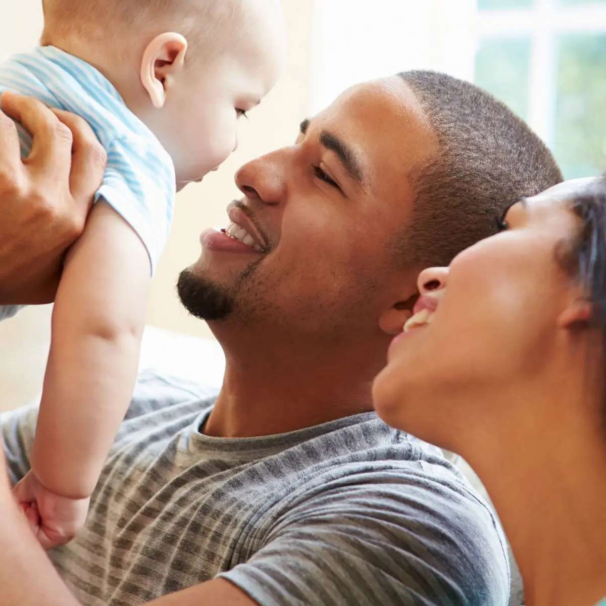 A man smiles while holding a baby up, and a woman sits beside them, also smiling. They appear happy and are indoors with natural light coming through a window.