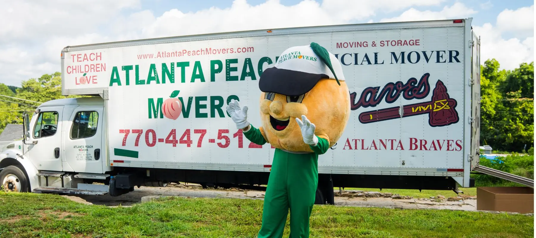 A person in a peach mascot costume stands waving in front of an Atlanta Peach Movers truck with Atlanta Braves branding, parked on grass under a partly cloudy sky.