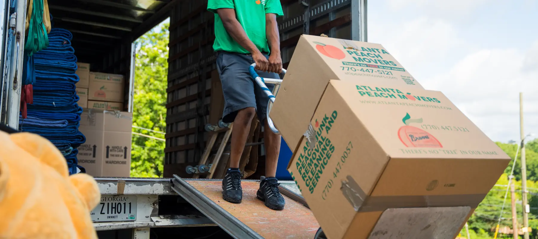 A person in a green shirt moves cardboard boxes labeled Atlanta Peach Movers on a dolly, unloading them from a truck parked outdoors.